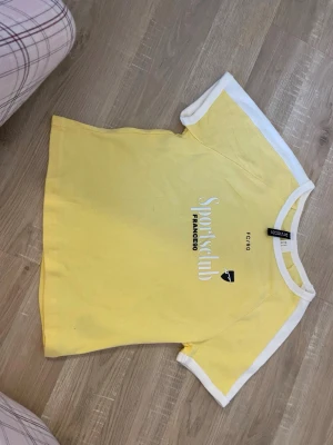 Gul och vit t-shirt från Fogg - Gul t-shirt från Fogg med vita kanter på ärmar och hals. Tryck på bröstet med texten 'Sportsclub FRANCE98'. Klassisk passform och kortärmad modell i mjuk bomull. Perfekt för en sportig och avslappnad stil. Mindre i storleken.