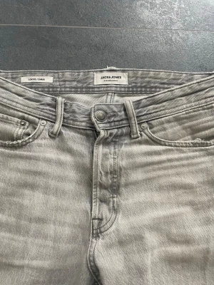 Grå vintage washed jeans från Jack & Jones - Gråa jeans från Jack & Jones i loose fit-modell. Klassisk femficksdesign med knappgylf och ljus tvättad denim. Snygga bakfickor med diskret broderi och läderpatch i midjan. Perfekta för en avslappnad och trendig stil.