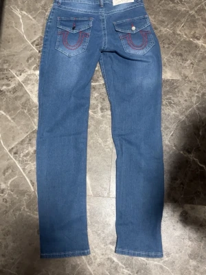 Blå True Religion jeans med röda detaljer - Snygga blå jeans från True Religion med raka ben och klassiska röda sömmar på bakfickorna. Jeansen har fem fickor och stängs med dragkedja och knapp. Perfekta för dig som gillar streetwear och vill sticka ut lite extra.