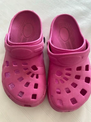 Rosa tofflor i crocs-stil - Säljer ett par rosa tofflor i klassisk crocs-stil med justerbar hälrem och ventilerande hål på ovansidan. Tillverkade i lätt och mjukt gummimaterial som är bekvämt att ha på sig. Perfekta för att glida runt hemma eller på stranden.