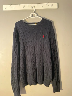 Mörkblå kabelstickad tröja Polo Ralph Lauren - Snygg mörkblå kabelstickad tröja från Polo Ralph Lauren med klassisk röd logga broderad på bröstet. Tröjan har rund halsringning, långa ärmar och ribbade muddar. Perfekt för dig som gillar tidlös och stilren design.