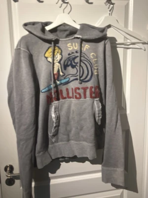Grå hoodie Hollister Surf Club - Grå hoodie från Hollister med broderad surfmotiv och texten 'Surf Club' på bröstet. Stor ficka framtill, dragsko i huvan och ribbade muddar. Skön och avslappnad stil med coolt tryck i flera färger. Perfekt för dig som gillar streetwear och surfvibbar.