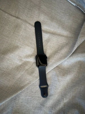 Svart Apple Watch med silikonarmband - Snygg svart Apple Watch med fyrkantig urtavla och svart silikonarmband. Klockan har en modern och stilren design med digital display och sensor på baksidan. Perfekt för dig som vill ha koll på både träning och notiser direkt på handleden. 2nd gen, använd i ca ett halvt år