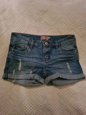 Blå jeansshorts med slitningar - Snygga blå jeansshorts från Miss Milla med slitna detaljer framtill och uppvikta benslut. Klassisk femficksmodell med knapp och dragkedja. Perfekta för varma dagar och enkel att matcha med allt. Säljer då de inte passar mig.