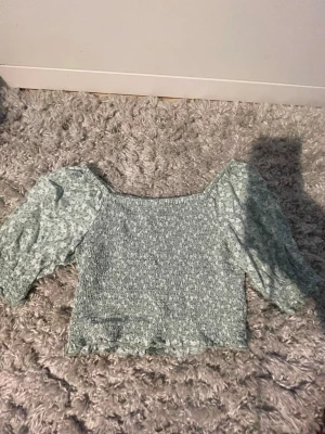 Grön blommig offshoulder blus - Söt ljusgrön blus med små vita blommor, fyrkantig urringning och puffiga korta ärmar. Volangkant nertill. Perfekt till jeans eller kjol för en somrig fin look