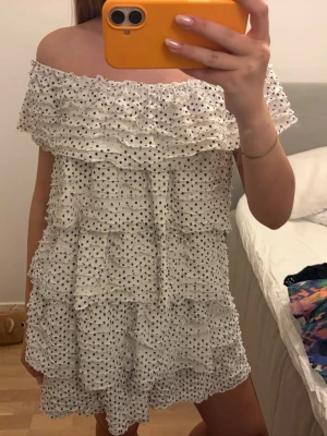 Vit offshoulder prickig kortklänning - Supersöt vit kortklänning med svarta prickar och volangdetaljer. Offshoulder-modell som ger en trendig vibe och luftig känsla. Perfekt för varma dagar och sommarhäng. Klänningen har flera lager volanger och är tillverkad i ett lätt material.