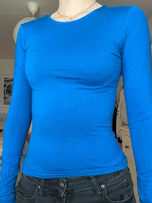 Kroppsnära blå långärmad topp - Snygg och enkel långärmad topp i klarblå färg med rund halsringning. Modellen är tight och följsam, perfekt att styla med jeans eller kjol. Materialet ser ut att vara stretchigt och mjukt, vilket gör den bekväm att bära hela dagen.
