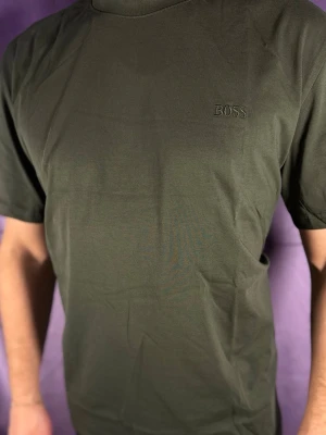 Olivgrön t-shirt från BOSS xl-xxl - Snygg olivgrön t-shirt från BOSS med diskret broderad logga på bröstet. Klassisk passform och rund halsringning, perfekt för en stilren och enkel look. Tillverkad i mjuk bomull som känns skön mot huden.