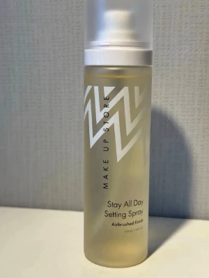 Make Up Store Stay All Day Setting Spray - Face mist från Make Up Store i genomskinlig plastflaska med vitt mönster och vit spraykork. Ger en airbrushed finish och håller sminket på plats hela dagen. Innehåller 100 ml. Perfekt för att fräscha upp och fixera din makeup.