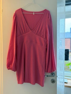 Rosa kort klänning med v-ringning - En rosa kort klänning med v-ringad hals och långa puffärmar. Klänningen har en lätt strukturerad yta och är tillverkad i ett mjukt material som ger en skön känsla. Perfekt för dig som gillar en feminin och trendig stil. Endast använd en kväll. 