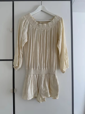 Krämvit boho jumpsuit Ralph Lauren  - Supersöt krämvit jumpsuit i boho-stil med långa ärmar och smockdetaljer vid halsringning och midja. Tillverkad i ett lätt, luftigt tyg med struktur. Perfekt för en avslappnad och trendig look.