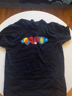 Svart t-shirt med boxningsmotiv - Svart t-shirt med tryck av boxningshandskar mellan två klubbmärken på bröstet. Klassisk passform och rund halsringning. Perfekt för dig som gillar sportiga och unika motiv.