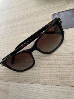 Tom Ford bruna solglasögon - Snygga solglasögon från Tom Ford med brunmelerad båge och bruna tonade glas. Klassisk fyrkantig form med diskret logga på sidan. Perfekt accessoar för att lyfta din stil.