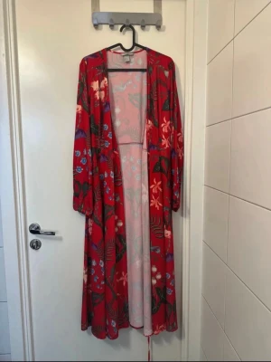 Röd blommig omlottklänning - Snygg röd omlottklänning med blommigt mönster och långa ärmar. Klänningen har v-ringning och knytband i midjan för en smickrande passform. Perfekt för dig som gillar färg och mönster.
