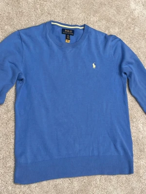 Ljusblå tröja från Polo Ralph Lauren - Snygg blå tröja från Polo Ralph Lauren med rund halsringning och klassisk gul broderad logga på . Perfekt för en stilren och avslappnad look. Tröjan är storlek 14/16 men är liten i storleken.