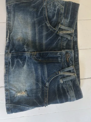 Blå jeanskjol med slitningar - Snygg blå jeanskjol i kort modell med slitna detaljer och klassiska fickor fram och bak. Kjolen har bälteshällor och knappstängning framtill. Vintage!