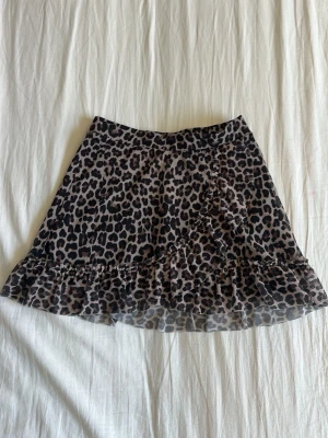 Leopardmönstrad volangkjol - Kort kjol med leopardmönster och volangkant. Kjolen har en lätt och luftig känsla, perfekt för dig som vill sticka ut. Färgerna går i beige, svart och brunt. Materialet känns som polyester och kjolen har en omlott-look med volangdetaljer.
