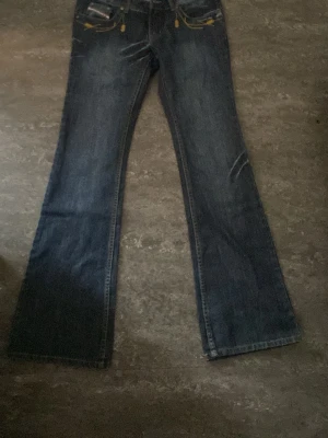 Mörkblå vintage bootcut jeans med brodyr - Säljer ett par mörkblå bootcut jeans med slitningar och coola broderade detaljer i gult på bakfickorna. Jeansen har en klassisk femficksmodell och synliga slitningar framtill för en trendig look. Jeansen är långa och passar mig som är 173 dom går till och med lite över. Jeansen är köpte från en hemsida som heter tinlessel. Pris kan diskuteras