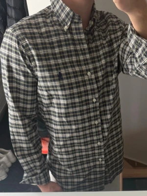 Rutig skjorta från Ralph Lauren - Klassisk långärmad skjorta från Ralph Lauren i blått, vitt och grönt rutigt mönster. Skjortan har button-down krage, knäppning framtill och broderad logga på bröstet. Perfekt för en stilren och avslappnad look.