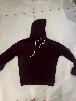 Vinröd hoodie - Mysig vinröd hoodie med klassisk huva och vita dragsnören. Hoodien har en enkel design utan tryck eller logga, samt en stor ficka framtill. Perfekt för chill dagar eller när du vill ha en clean look. Tillverkad i mjukt material för extra komfort.
