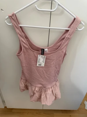 Rosa volangtopp från H&M DIVIDED - Supersöt ljusrosa topp från H&M DIVIDED med volang nertill. Ärmlös modell med rundad halsringning och mjukt bomullstyg. Perfekt för en trendig och avslappnad look.
