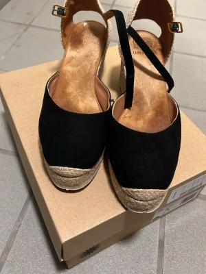 Svarta espadrille-sandaler Unisa - Snygga svarta espadrille-sandaler från Unisa med kilklack i flätad jute och justerbar rem runt vristen. Ovandelen är i mockaliknande material och sulan har en klassisk espadrille-look. Perfekta för sommaren och ger en trendig touch till din outfit.
