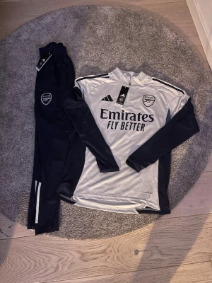 Arsenal träningsset från Adidas - Snyggt Arsenal träningsset från Adidas med långärmad tröja och byxor. Tröjan är vit med svarta ärmar och detaljer, har halv dragkedja och klubbmärke på bröstet. Byxorna är svarta med vita detaljer och Arsenal-logga. Materialet är lätt och andas, perfekt för träning.