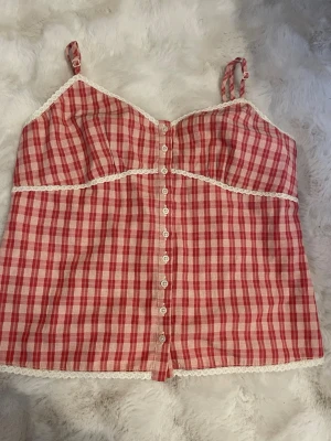 Rutig röd linnetopp Brandy Melville - Supersöt rutig linnetopp från Brandy Melville i rött och vitt. Toppen har tunna axelband, knappar framtill och spetsdetaljer längs kanterna. Perfekt för varma dagar och ger en vintagevibe till din outfit.