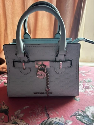 Ljusblå handväska från Michael Kors - Snygg ljusblå handväska från Michael Kors med präglat MK-mönster, dubbla handtag och silverfärgade metalldetaljer. Väskan har ett dekorativt hänglås framtill och en kedjedetalj med MK-logga. Perfekt accessoar för att lyfta din outfit. Aldrig använd och Äkta såklart! Nypris är ca €395
