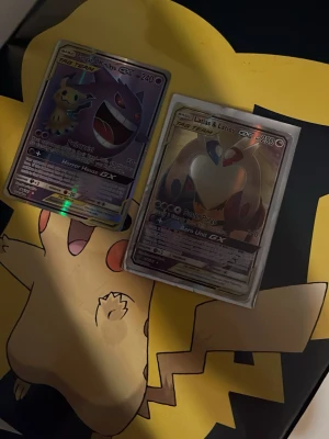 Pokémon GX-kort i plastfickor - Två Pokémon GX-samlarbilder i plastfickor, en med Gengar & Mimikyu och en med Latias & Latios. Kortens design är färgstark med holografiska effekter och detaljerade illustrationer. Perfekt för dig som gillar Pokémon och samlarkort.