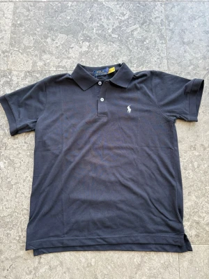 Svart pikétröja från Ralph Lauren - Klassisk svart pikétröja från Ralph Lauren med vit broderad logga på bröstet. Tröjan har korta ärmar, krage och två knappar framtill. Perfekt för en stilren och avslappnad look. Viktigt att veta är att den är liten i st, den passar även folk som brukar ha M!