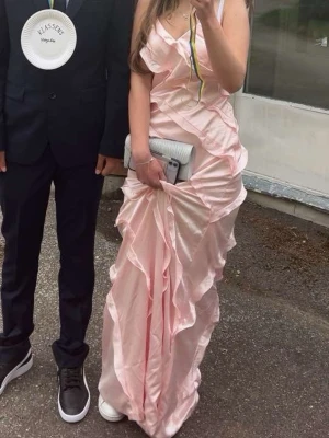 Ljusrosa långklänning med volanger - Säljer en elegant ljusrosa långklänning med volangdetaljer och omlottkänsla. Klänningen har smala axelband och en feminin, figurnära passform. Perfekt för speciella tillfällen när du vill sticka ut med en romantisk look. Balklänning använd 1 gång 