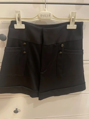 Svarta shorts från Zara  med knappar - Stilrena svarta shorts från Zara med hög midja och dekorativa guldknappar framtill. Shortsen har stora fickor på sidorna och är tillverkade i ett mjukt, lite stretchigt material som ger en snygg passform. Perfekta för en trendig och avslappnad look.
