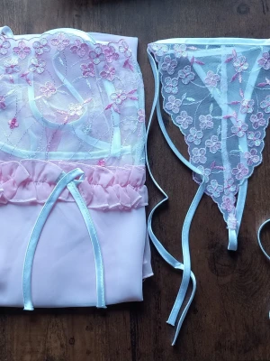 Rosa spetsbody med string från Enigma storlek S - Sött set från Enigma med en ljusrosa body i satin och spets, dekorerad med blommig brodyr och tunna axelband. Matchande stringtrosa i transparent spets med blommor och smala band. Perfekt för dig som gillar romantiska detaljer. Storlek S