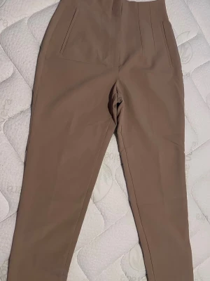 Beige höga kostymbyxor - Snygga beige kostymbyxor med hög midja och raka ben. Byxorna har diskreta fickor fram och stilrena sömmar. Perfekta för en clean och modern look. Materialet känns mjukt och följsamt, vilket ger en bekväm passform.