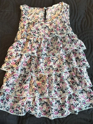 Blommig volangblus i pastellfärger - Supersöt blommig blus med flera volanglager och ärmlös design. Mönstret går i rosa, svart och vit med små blommor över hela toppen. Perfekt för dig som gillar romantisk och lekfull stil.