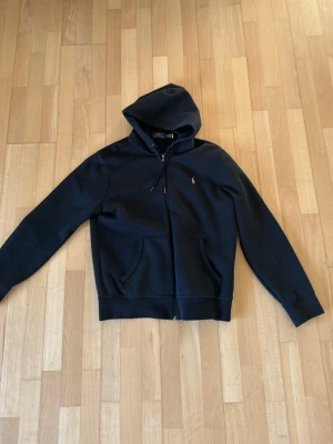 Svart hoodie  Ralph Lauren - Svart hoodie med dragkedja från Ralph Lauren. Klassisk design med huva, två fickor framtill och den ikoniska broderade loggan på bröstet. Perfekt för en avslappnad och stilren look. Mjuk bomullsblandning för extra komfort.