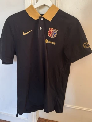 FC Barcelona pikétröja Nike - Svart pikétröja från Nike med FC Barcelona-logga och Spotify-sponsor på bröstet. Tröjan har gul krage, korta ärmar och knappar framtill. Perfekt för dig som älskar fotboll och vill visa ditt stöd för Barça i sommer