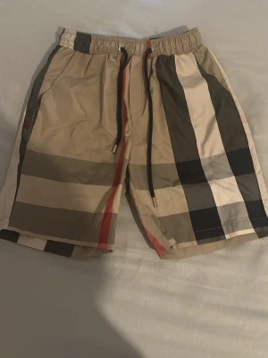 Beige rutiga shorts med snörning - Snygga beige shorts från Burberry med klassiskt rutigt mönster i svart, vitt och rött. Shortsen har elastisk midja med snörning och sidofickor. Perfekta för sommaren och ger en stilren look.