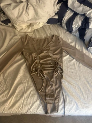 Beige ribbad body med snörning - Snygg beige body med långa ärmar och ribbad struktur. Modellen har snörning framtill som ger en cool detalj och tryckknappar i grenen för enkel på- och avtagning. Perfekt att styla med jeans eller kjol för en trendig look. 