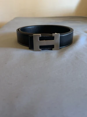 Svart märkes bälte  - Säljer denna svarta märkesbältet med silver buckla och vita stygn längs kanterna. Stilig och fräsch, perfekt för en simpel outfit!