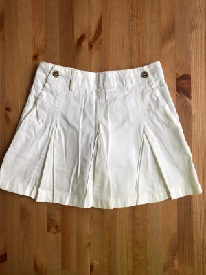 Vit plisserad skort med fickor CROSS - Snygg vit skort med plisserade detaljer framtill och två knappar i midjan. Skorten har bälteshällor och fickor både fram och bak. Perfekt för en fräsch och stilren look. Tillverkad i ett mjukt bomullstyg.