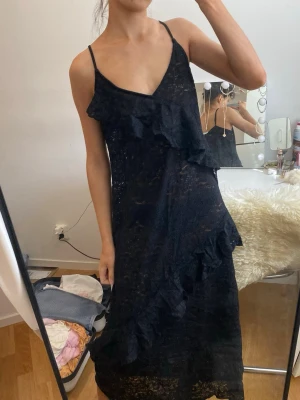 Svart volangklänning med spetsdetaljer - Snygg svart långklänning med tunna axelband och volangdetaljer framtill och längs kjolen. Klänningen har spetsmaterial som ger en elegant känsla och en feminin siluett. Perfekt för dig som vill sticka ut med en stilren look.
