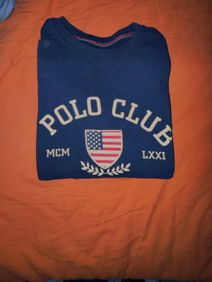 Blå sweatshirt från Vinson Polo Club - Mörkblå sweatshirt från Vinson Polo Club med stor vit text och amerikansk flagga på bröstet. Rund halsringning med röd detaljsöm på insidan. Tillverkad i mjukt material, perfekt för en avslappnad och sportig stil.