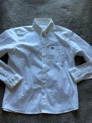 Vit skjorta från Lacoste - Klassisk vit skjorta från Lacoste med button-down krage och bröstficka med den ikoniska krokodilloggan. Skjortan är långärmad och tillverkad i mjuk bomull, perfekt för en clean och stilren look.