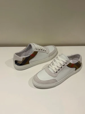 Burberry vita sneakers med rutigt mönster - Snygga vita sneakers från Burberry med klassiskt rutigt mönster i beige, svart och rött på sidorna. Skorna har vita snören, detaljer i mocka och en stilren låg profil. Perfekta för dig som vill ha en clean look med en touch av lyx.