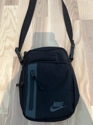 Nike svart axelväska med logga - Snygg svart axelväska från Nike med justerbar rem och dragkedjefack framtill. Väskan har en stilren design med grå detalj och Nike-logga på framsidan. Perfekt för att bära dina viktigaste grejer.
