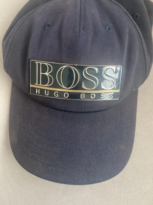 Mörkblå keps från Hugo Boss - Snygg mörkblå keps från Hugo Boss med stor metallbricka framtill där det står BOSS. Klassisk böjd skärm och justerbar passform. Perfekt accessoar för en stilren och sportig look.