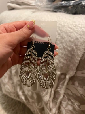 Guldiga fjäderörhängen med stenar - Snygga örhängen i form av fjädrar från Accessories. De är guldiga med glittrande stenar och en droppformad detalj i mitten. Perfekta för att ge din look lite extra bling. Lätta att bära och riktigt trendiga.