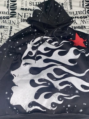 Hellstar hoodie  - Unik svart hoodie med stor vit grafisk illustration av ett ansikte och eldliknande mönster, samt en röd stjärna på bröstet. Hoodien har huva med dragsko och tryckta detaljer över hela plagget. Perfekt för dig som gillar streetwear och statement-plagg.